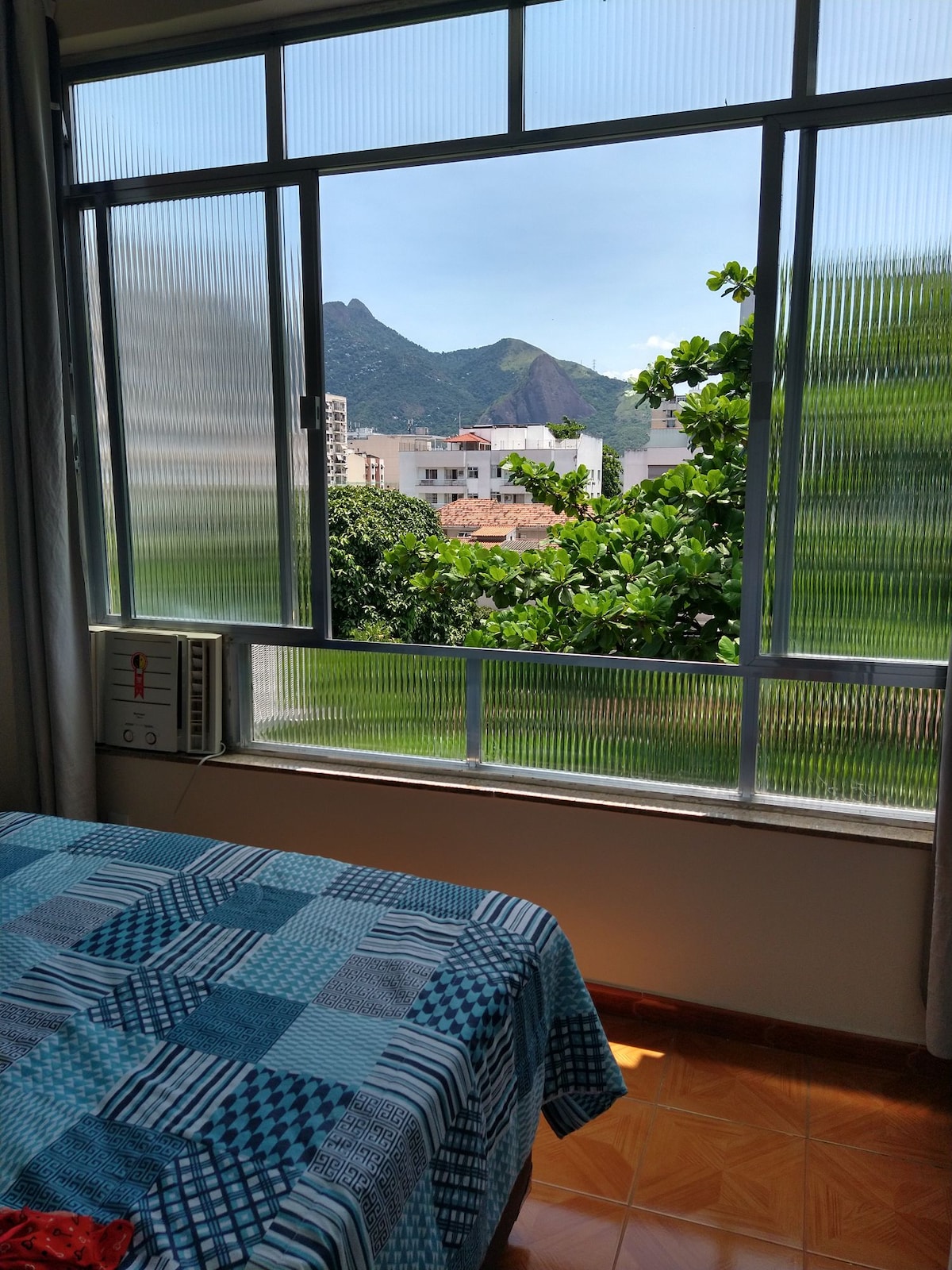 A bright bedroom features a large window offering views of surrounding greenery and mountains. Soft light illuminates a bed covered with a patterned blue blanket. Air conditioning is visible in the corner, ensuring comfort during warm days.