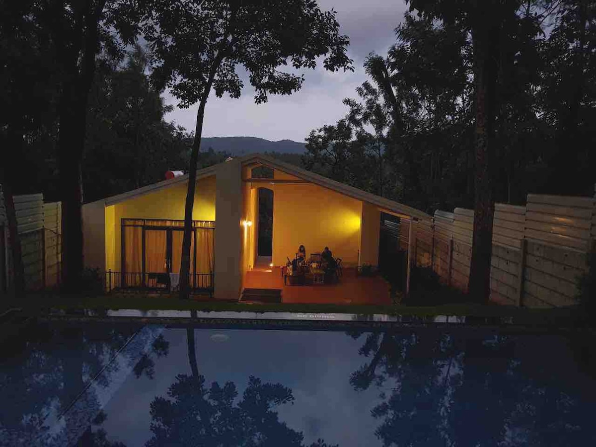 A serene evening view captures the property illuminated by warm lighting, with a swimming pool reflecting the scenery. Two individuals are seen on a porch area, surrounded by lush trees, with the distant hills creating a peaceful backdrop.