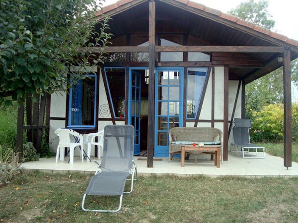 A charming exterior of a bungalow features wooden beams and large blue-framed glass doors. A small seating area with a cozy sofa is visible, along with two lounge chairs positioned on the patio. Lush greenery surrounds the entrance, enhancing the inviting atmosphere.