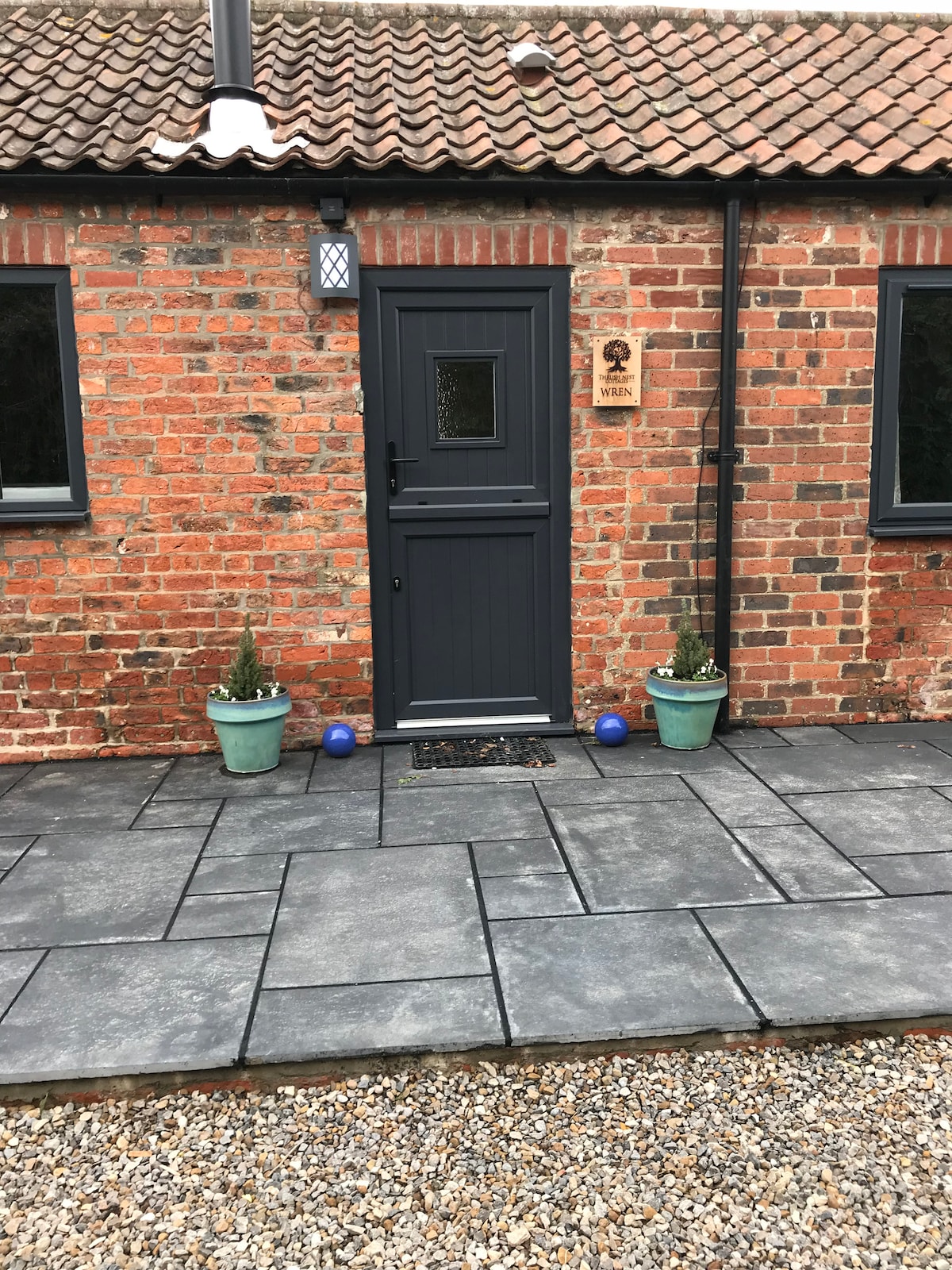 The entrance to a rustic cottage is showcased, featuring a dark gray door framed by rustic red brick. Two potted plants are positioned on either side of the door, and a smooth stone pathway leads to the entrance, complementing the gravel surface.
