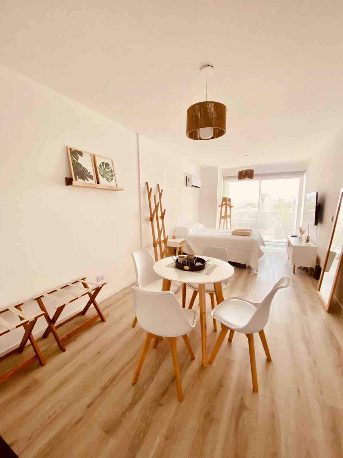 A light-filled dining area is presented, featuring a round table surrounded by four white chairs. A minimalist wooden rack stands nearby, with decorative wall art displayed on the adjacent wall. The spacious living area is visible in the background, complemented by a large window.