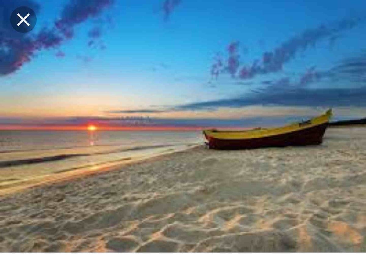 An empty beach scene features a wooden boat with a yellow hull resting on soft, sandy shores. The water reflects the shades of a sunset, with vibrant blues and warm oranges blending into the sky, creating a serene coastal atmosphere.