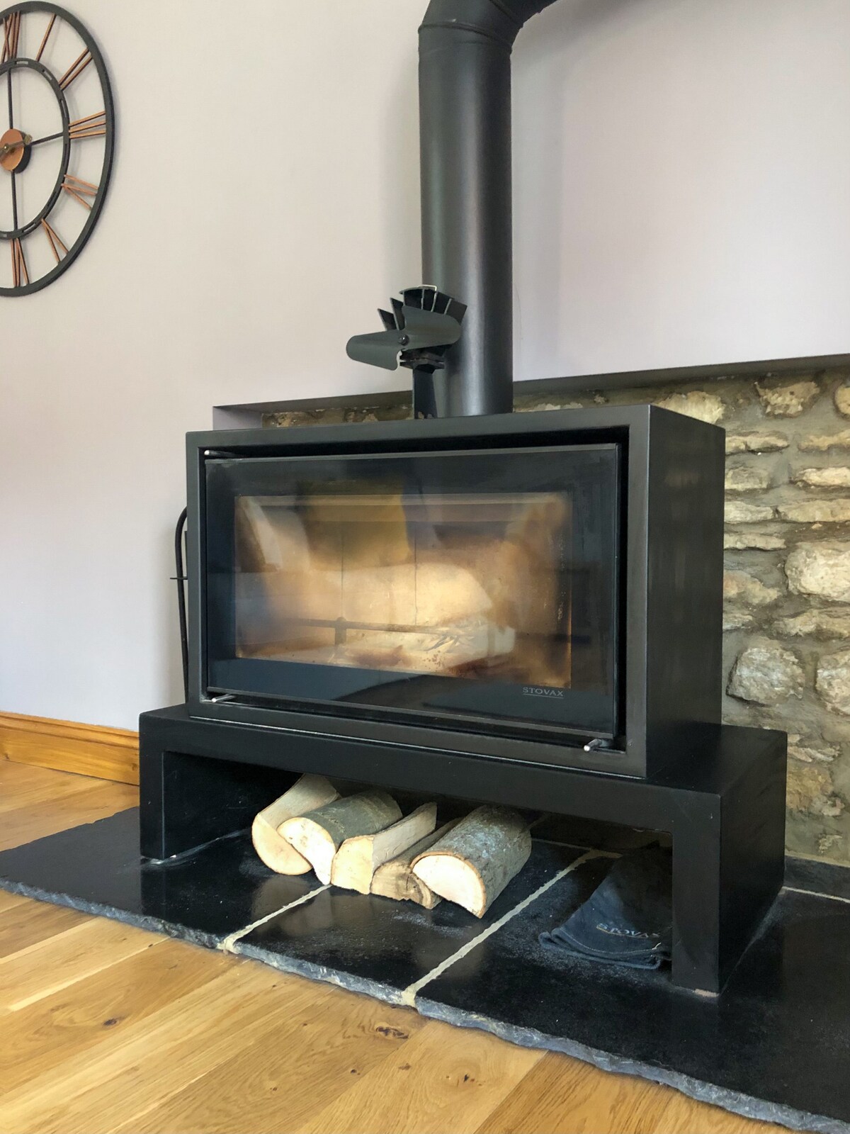 A modern log burner is situated against a stone wall, providing a rustic touch to the living area. Logs are neatly stacked beneath the unit, and a clock hangs on the wall above. The wooden floor enhances the warm, inviting ambiance.
