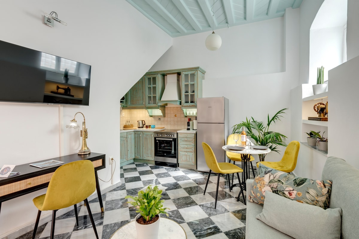 A bright kitchen area features muted teal cabinetry and a stainless steel refrigerator. A table with two chairs is paired with a small workspace. A cozy seating arrangement includes a light-colored sofa adorned with decorative pillows, while indoor plants add a touch of greenery.