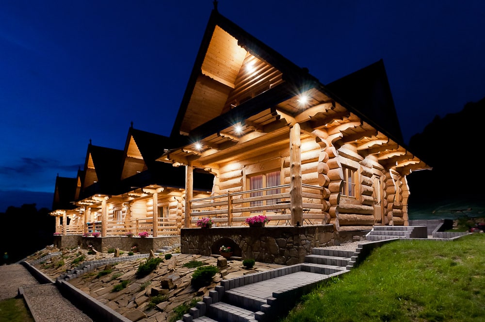 A row of rustic log cabins is illuminated against a twilight sky, showcasing wooden beams and stone accents. Each cabin features a spacious porch adorned with flowers, while a stone path leads to the entrance, enhancing the serene mountain surroundings.