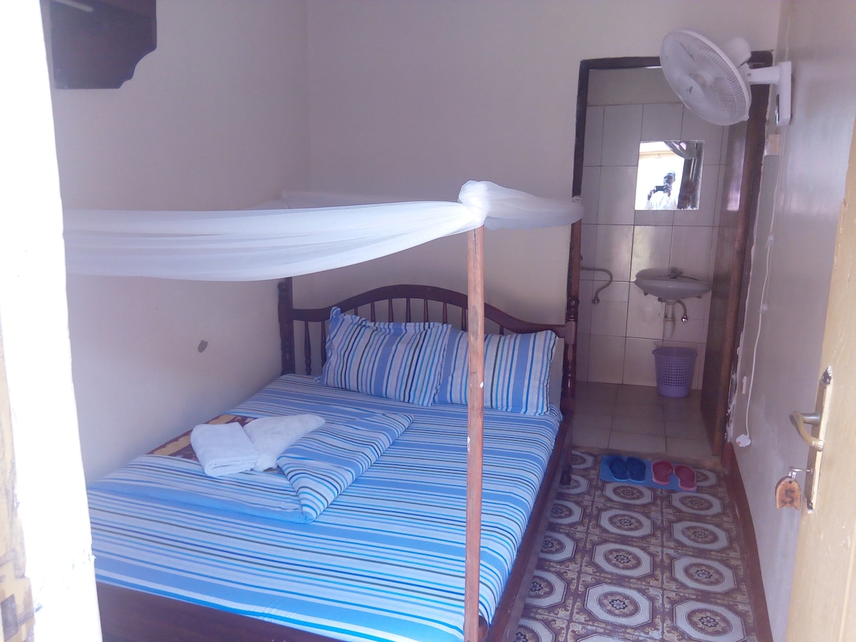 A well-furnished bedroom features a spacious bed dressed in blue and white striped bedding. A white mosquito net is suspended above the bed. A small sink and towel are visible in the corner, along with a pair of slippers on the tiled floor.