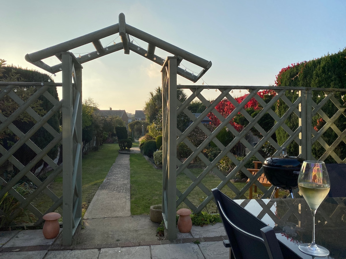 A garden path leads through a wooden trellis archway, framed by neatly trimmed hedges and colorful flowerbeds. A dining table and chairs are visible in the foreground, with a glass of wine placed on the table, reflecting a tranquil evening atmosphere.