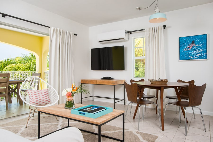 A bright living area includes a large flat-screen TV mounted on the wall, with a wooden dining table accompanied by four chairs. Natural light enters through windows adorned with white curtains, and a decorative coffee table sits in the center, complemented by colorful accents.