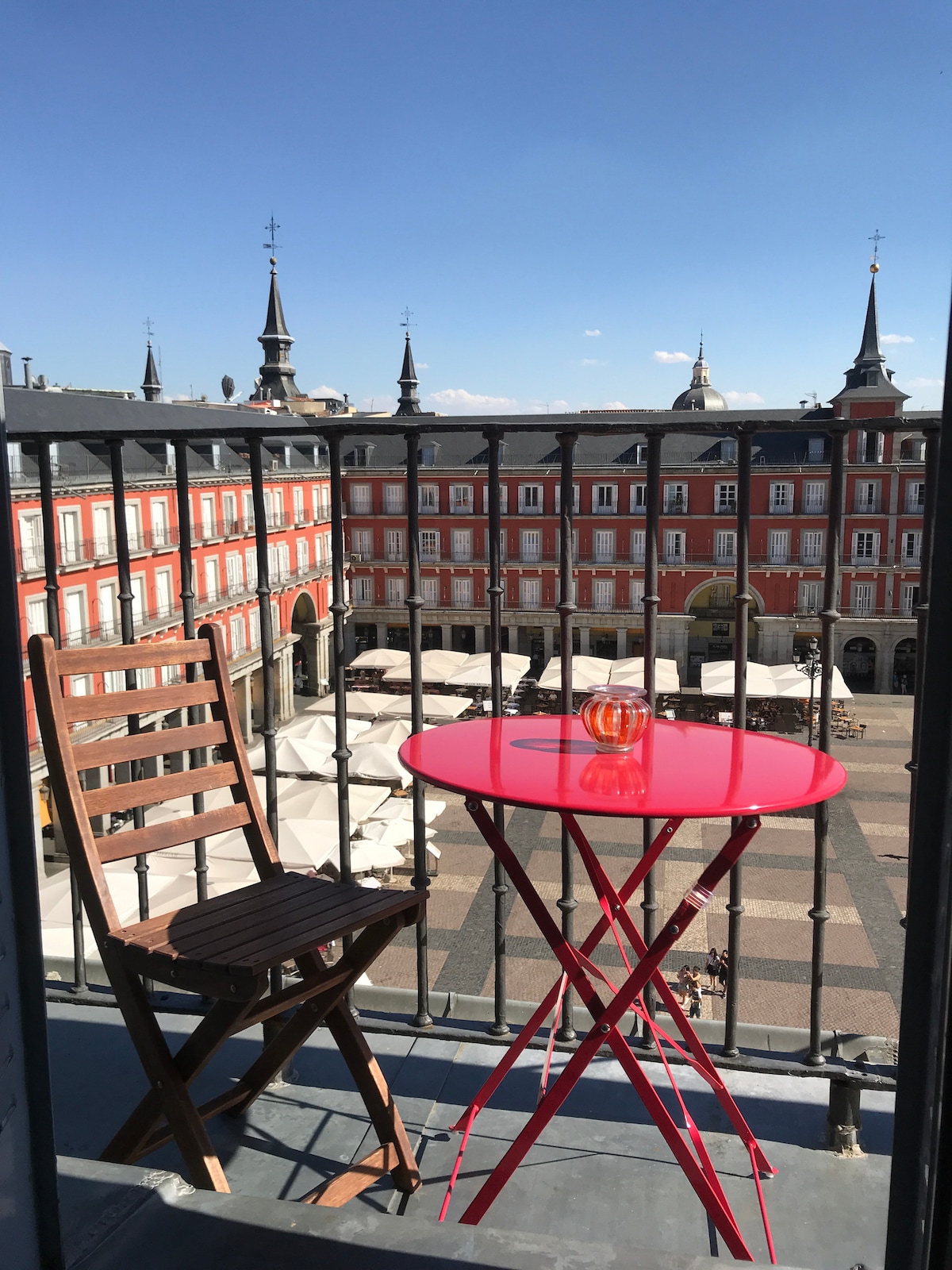 A small balcony offers a striking view of the Plaza Mayor, surrounded by historic buildings. A bright red table and a wooden chair are positioned on the balcony, with an elegant drink placed on the table. The clear blue sky enhances the vibrant ambiance.