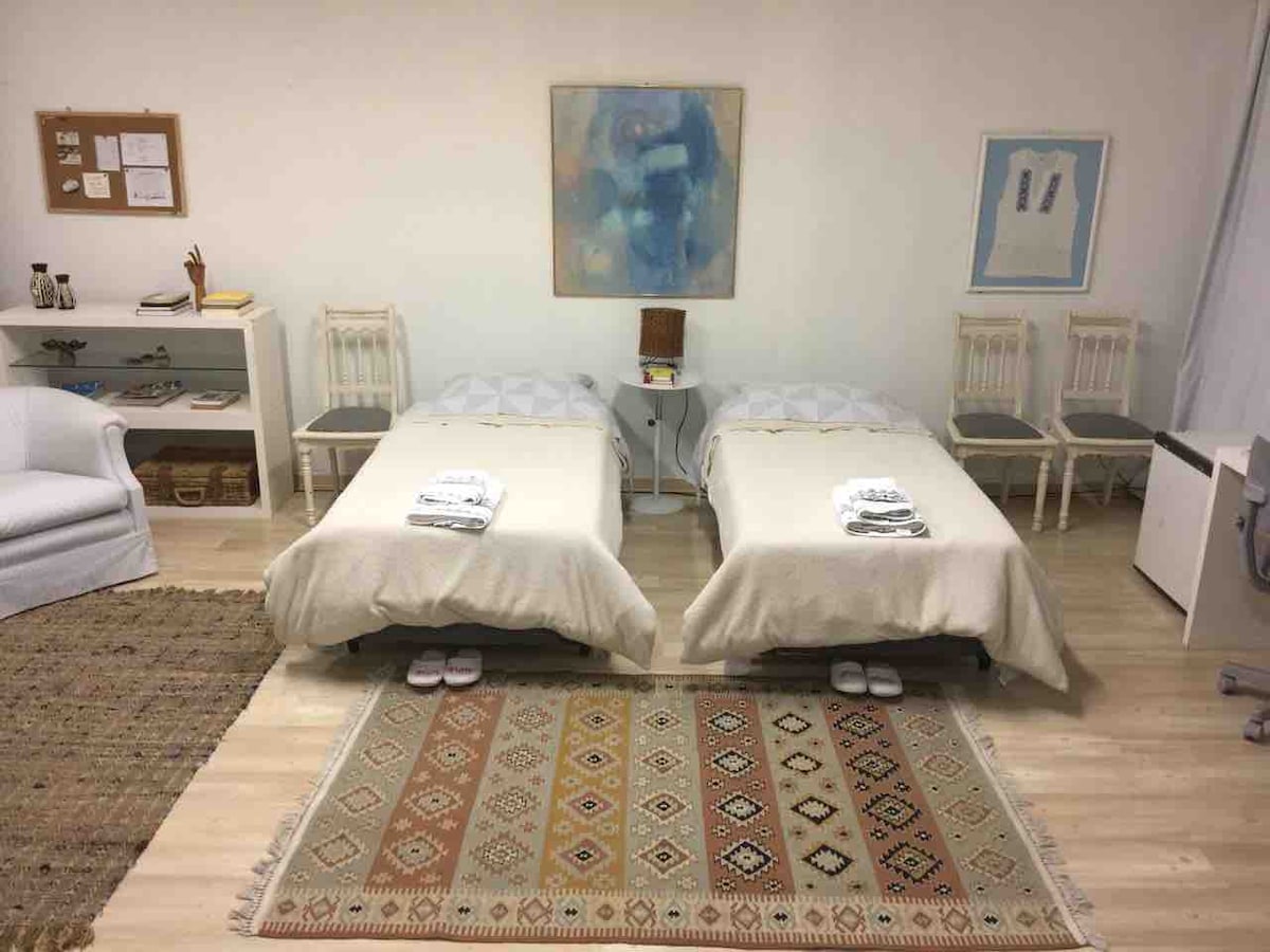 Two cozy beds with light bedding are positioned in the open space, complemented by a textured area rug. Simple wooden chairs and a white sofa enhance the room's aesthetic, while art pieces adorn the walls, contributing to a cultured ambiance.