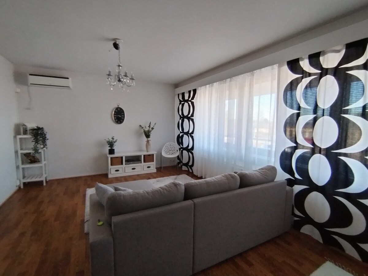 A light-filled living area features a large, comfortable sectional sofa facing a modern media unit. Decorative elements include a wall clock and potted plants. Floor-to-ceiling windows are adorned with bold black and white curtains, enhancing the spacious feel of the room.