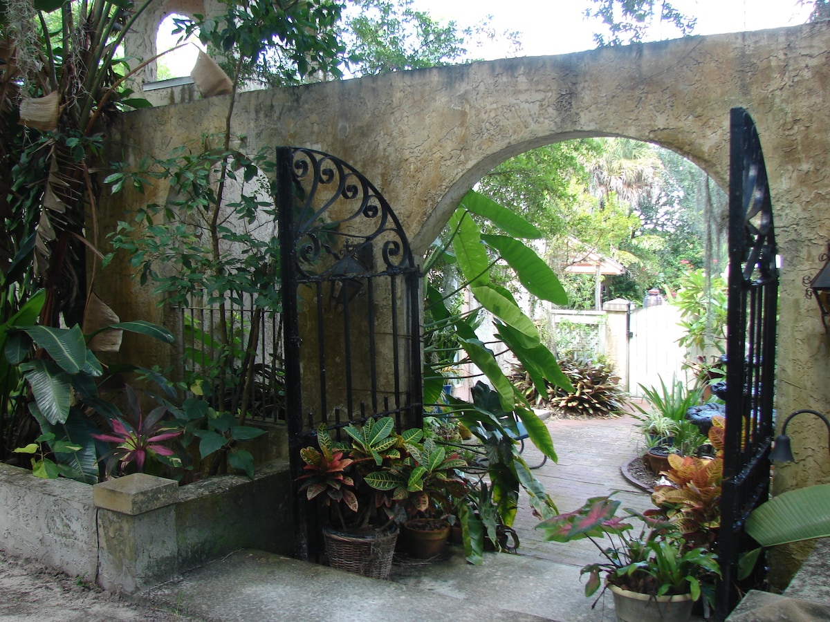 A wrought-iron gate opens into a lush courtyard, framed by a weathered stone wall. Various tropical plants are arranged in pots, showcasing vibrant greens and colorful foliage. The scene is serene, indicating an inviting and peaceful outdoor space within the estate.