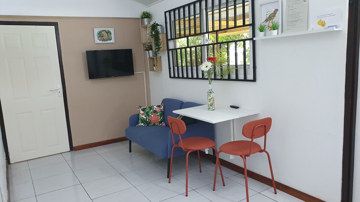 A small seating area is featured, with a blue couch positioned against the wall. A white table is accompanied by two orange chairs. Decorative plants and a vase with flowers are present on the table, creating a fresh and welcoming atmosphere. A wall-mounted TV is also visible.