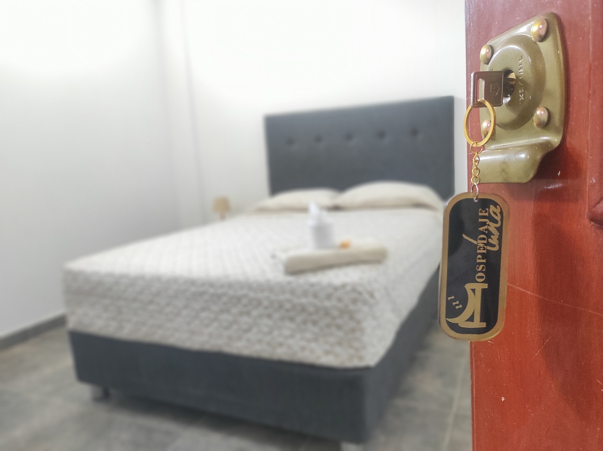 A spacious bed with a textured light-colored blanket is positioned against a gray upholstered headboard. A key with a decorative tag hangs from the door lock, suggesting a welcoming entrance to the private space.
