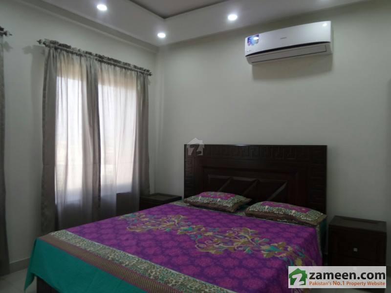 A well-lit bedroom features a spacious bed adorned with a vibrant purple and green blanket. Soft gray curtains frame a window, allowing natural light to brighten the room. An air conditioning unit is mounted above the bed, providing comfortable climate control.