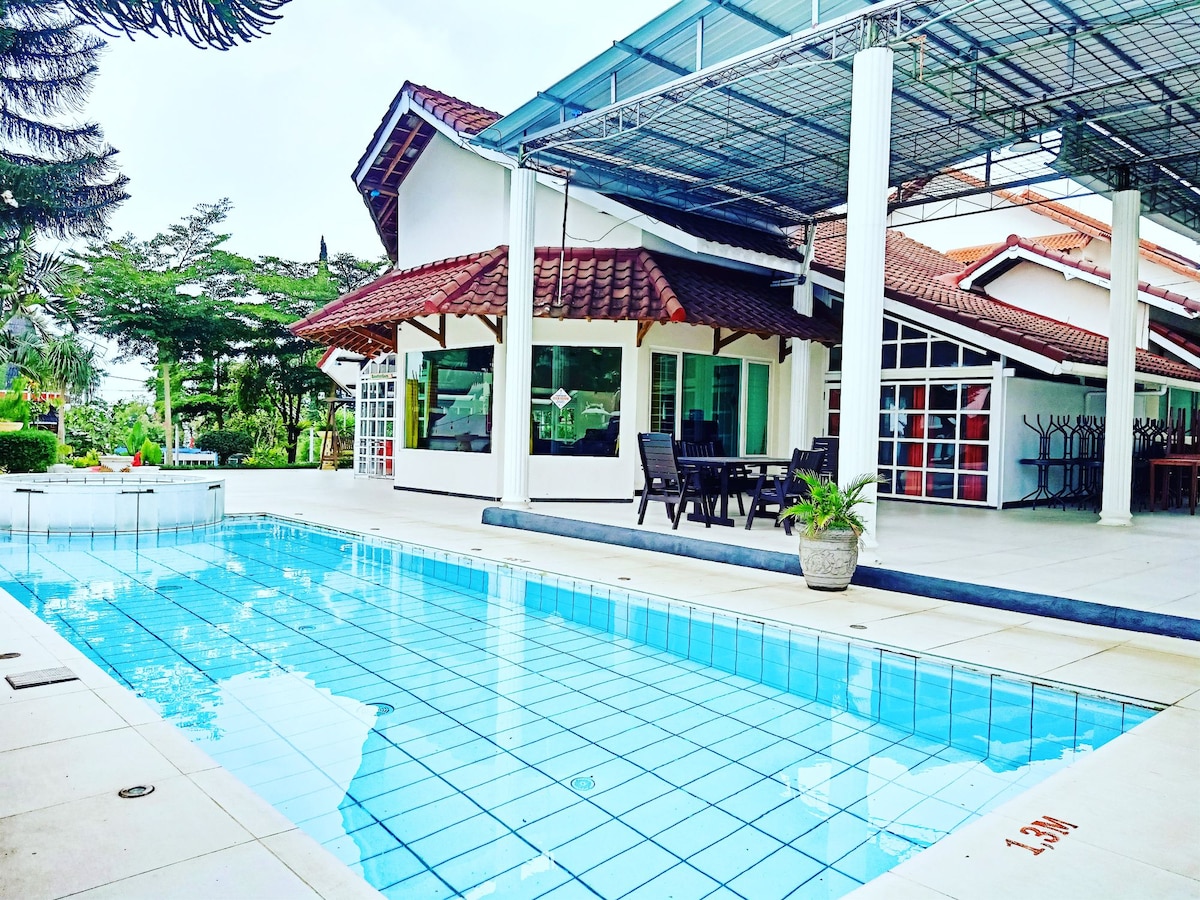 A well-maintained swimming pool is surrounded by a spacious patio area, featuring lounge chairs and outdoor dining furniture. The villa, with a sloped roof and large glass windows, provides a bright and open atmosphere, enhanced by lush greenery in the background.
