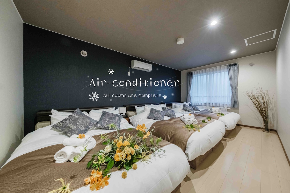 A spacious sleeping area features multiple beds, each dressed in crisp linens and decorative pillows. A large window allows natural light to fill the room, and an air-conditioning unit is mounted on the wall. A decorative arrangement of flowers adds a touch of warmth.