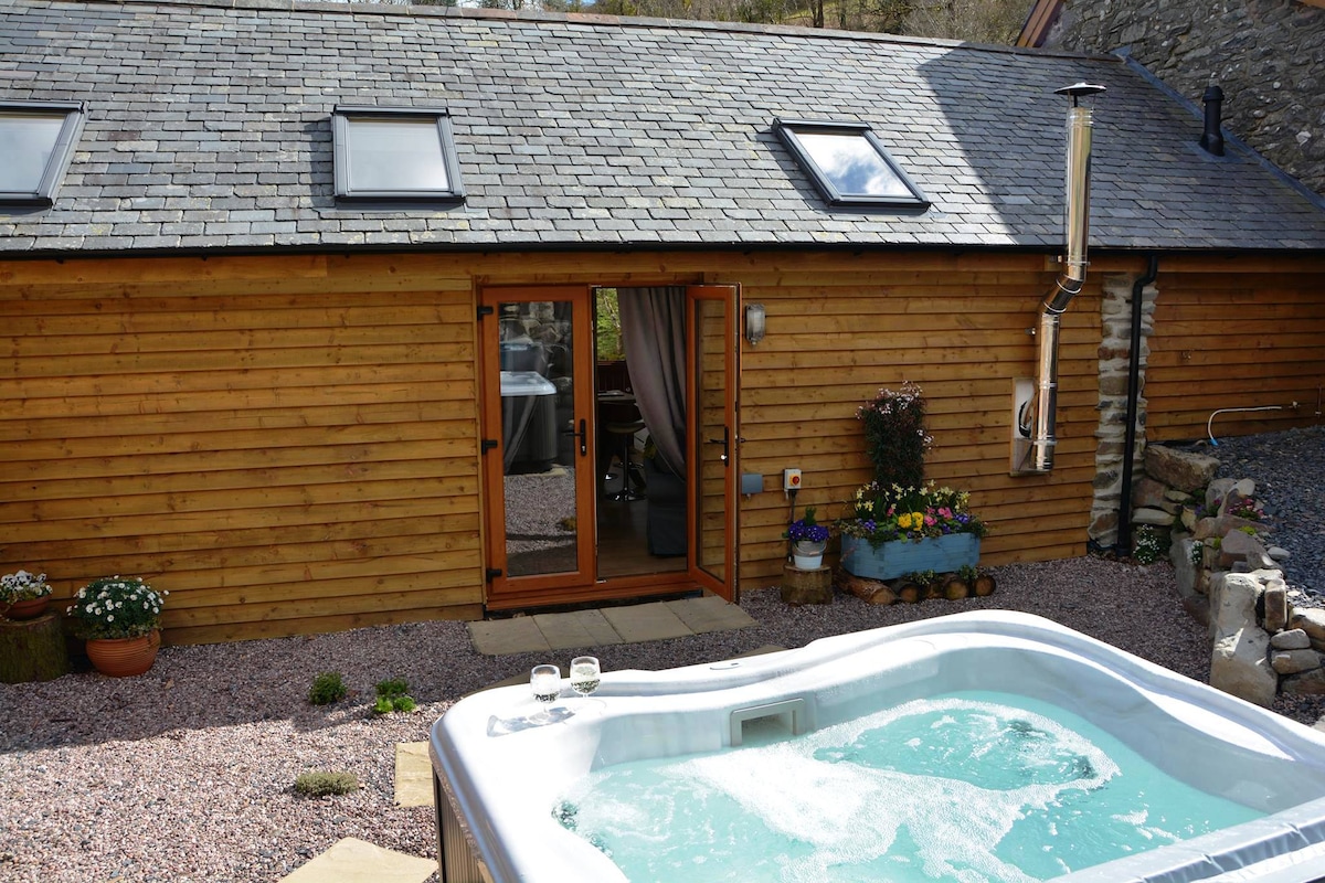 An outdoor area features a private hot tub, surrounded by gravel and flowers. French doors lead into a wooden-clad building, allowing a glimpse of the interior. Skylights are visible on the roof, enhancing the natural light entering the space.