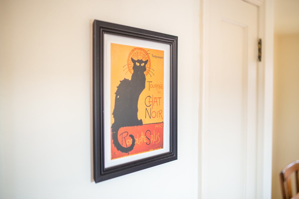A framed artwork depicting a stylized black cat against an orange background is displayed on a light-colored wall. The graphic features the text 'Tournee du Chat Noir' at the top, adding a touch of vintage charm to the space.