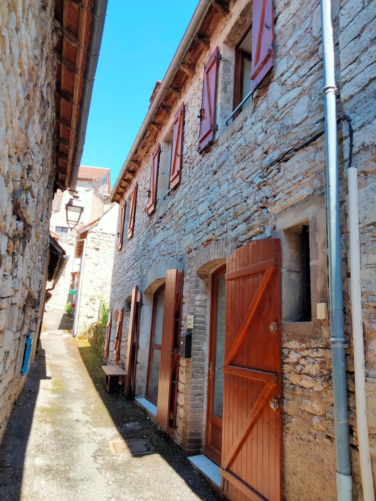A narrow, sunlit alleyway is flanked by stone buildings, showcasing wooden shutters and doors. The weathered stone walls exhibit a blend of textures. A small bench is positioned against one of the walls, inviting exploration of the charming surroundings.
