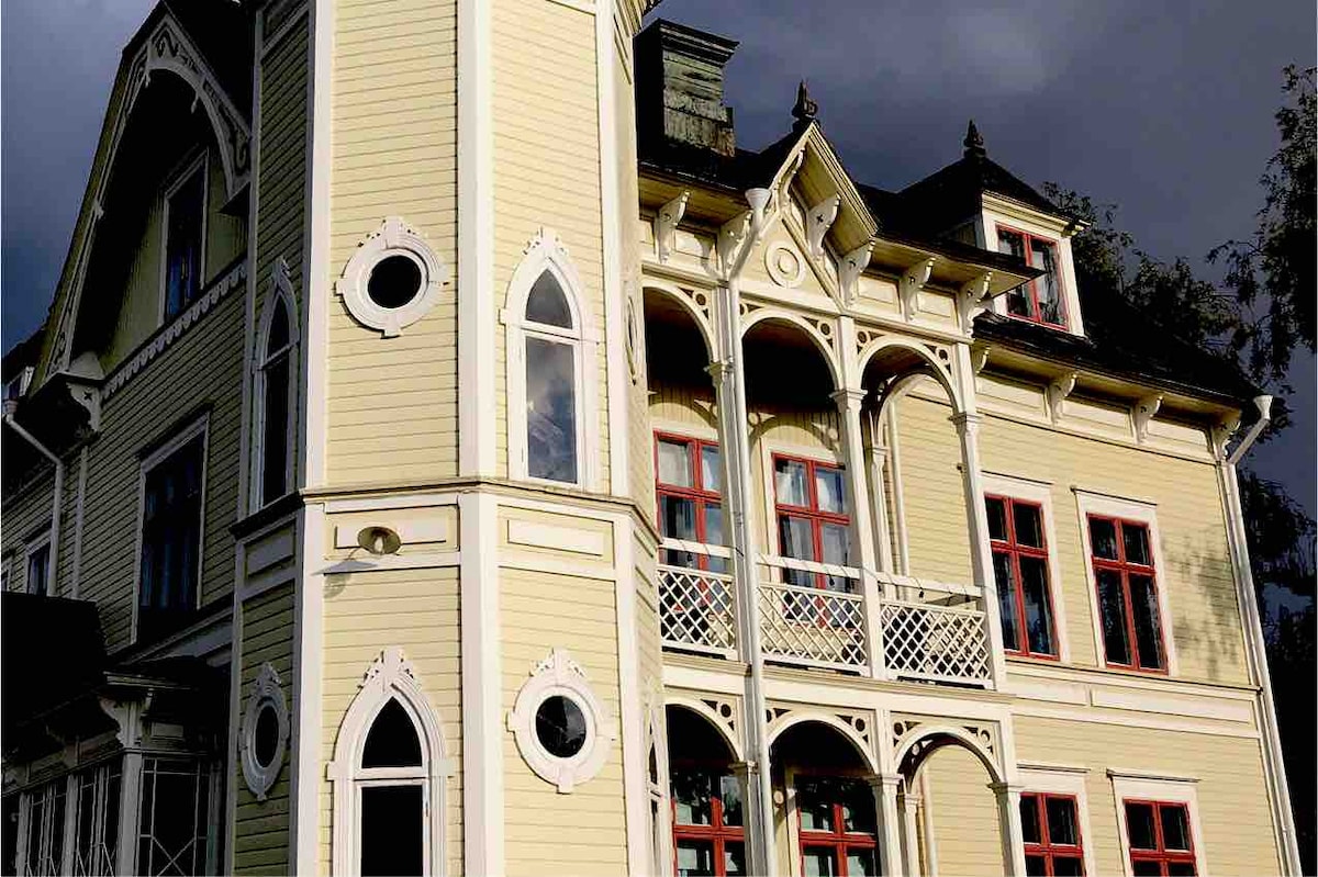 The exterior of an elegant house is showcased, featuring a distinctive architectural style with pointed arches and decorative trim. Large windows with red frames complement the light yellow siding, while intricate details enhance the overall charm of the structure.