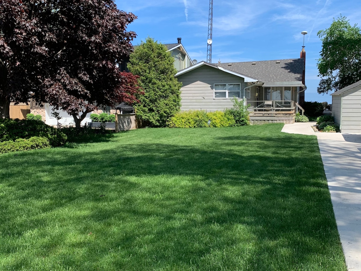 A spacious, well-maintained lawn is surrounded by a variety of trees and shrubs. The gray cottage is visible in the background, showcasing a welcoming facade. A concrete driveway leads up to the house, providing convenient access.