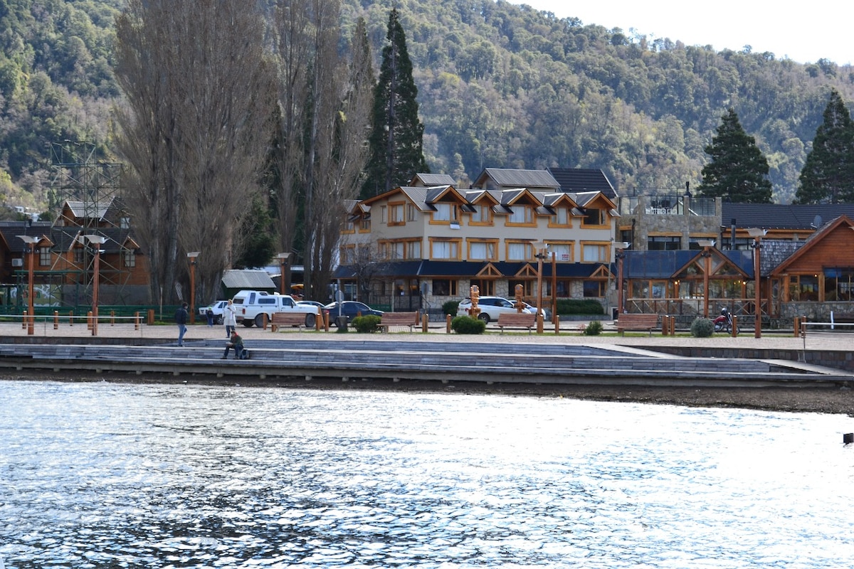 A lakeside view reveals a row of buildings characterized by wooden designs, surrounded by trees and mountainous terrain. A pathway along the water's edge features visitors enjoying the scenery, while the calm lake reflects the clear sky.