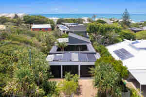 An aerial view of a modern home surrounded by lush greenery, with solar panels on the roof. The ocean is visible in the background, framed by the trees, while nearby houses also feature greenery and light-colored exteriors.