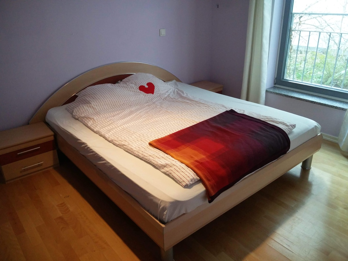 A spacious bed is positioned against a light purple wall, adorned with a decorative heart-shaped pillow. A soft blanket in warm tones lies across the foot of the bed. Natural light enters through a window, complemented by light-colored curtains.