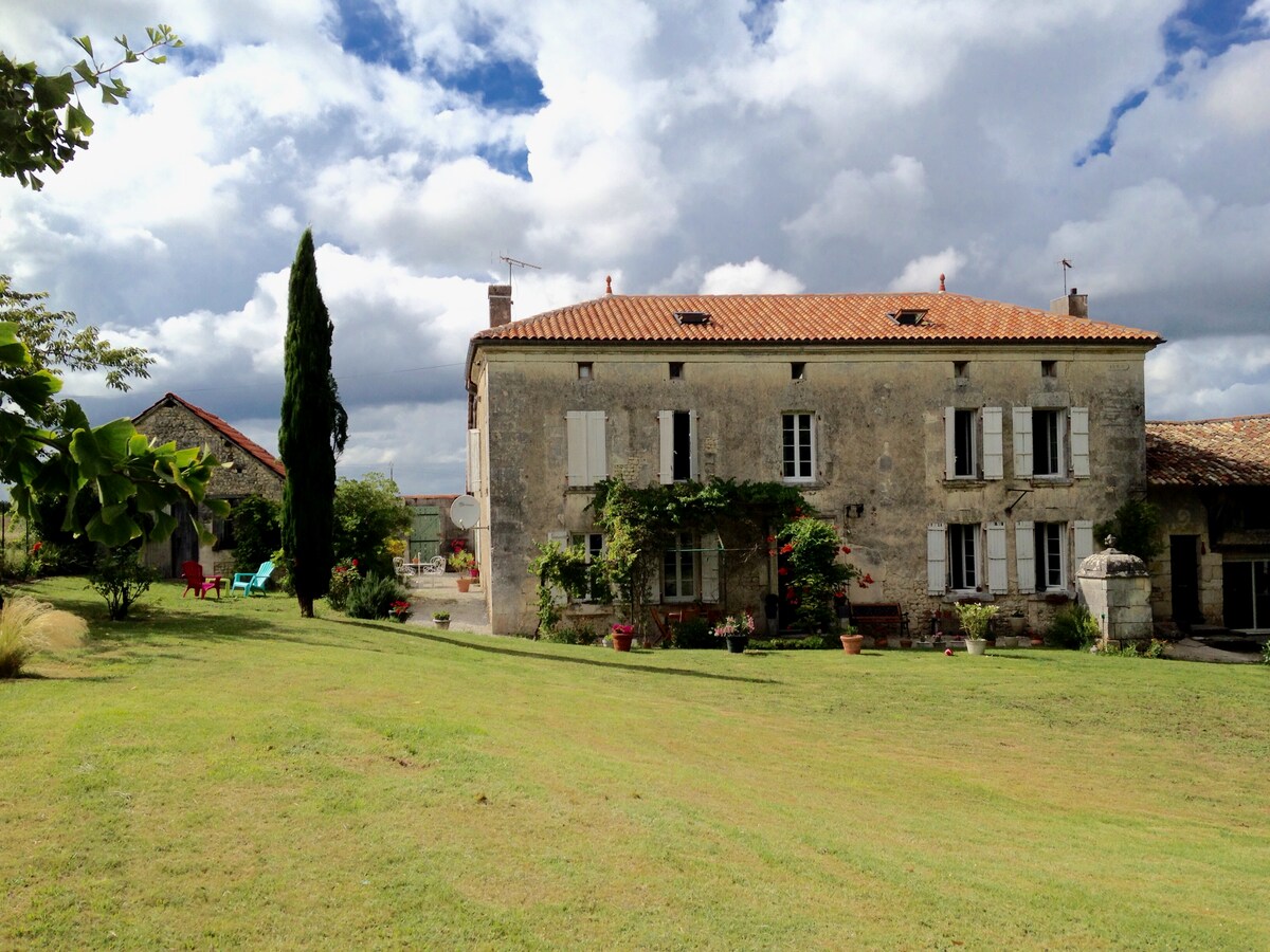 The charming Charentaise house is framed by lush greenery and a spacious lawn. Decorative shutters adorn the windows, while climbing plants add a touch of nature. A blue sky with fluffy clouds provides an inviting backdrop, showcasing the tranquil rural setting.