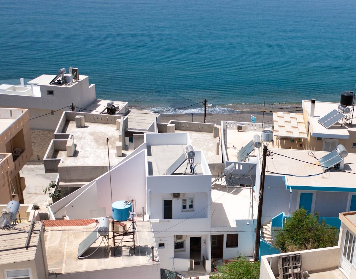 The image captures a panoramic view of coastal buildings with white rooftops, overlooking a tranquil sea. The blue waters reflect sunlight, while several water tanks and satellite dishes are visible on the rooftops, contributing to the local architecture.
