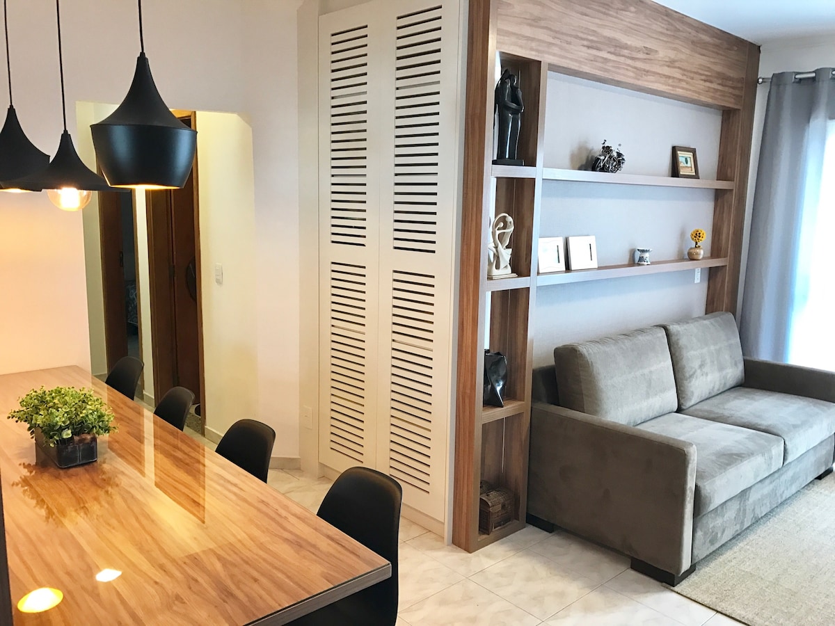 The dining area features a wooden table surrounded by black chairs, illuminated by stylish pendant lights. A cozy seating area with a gray sofa is positioned next to a wooden shelf displaying decorative items. Soft natural light enters through a large window.