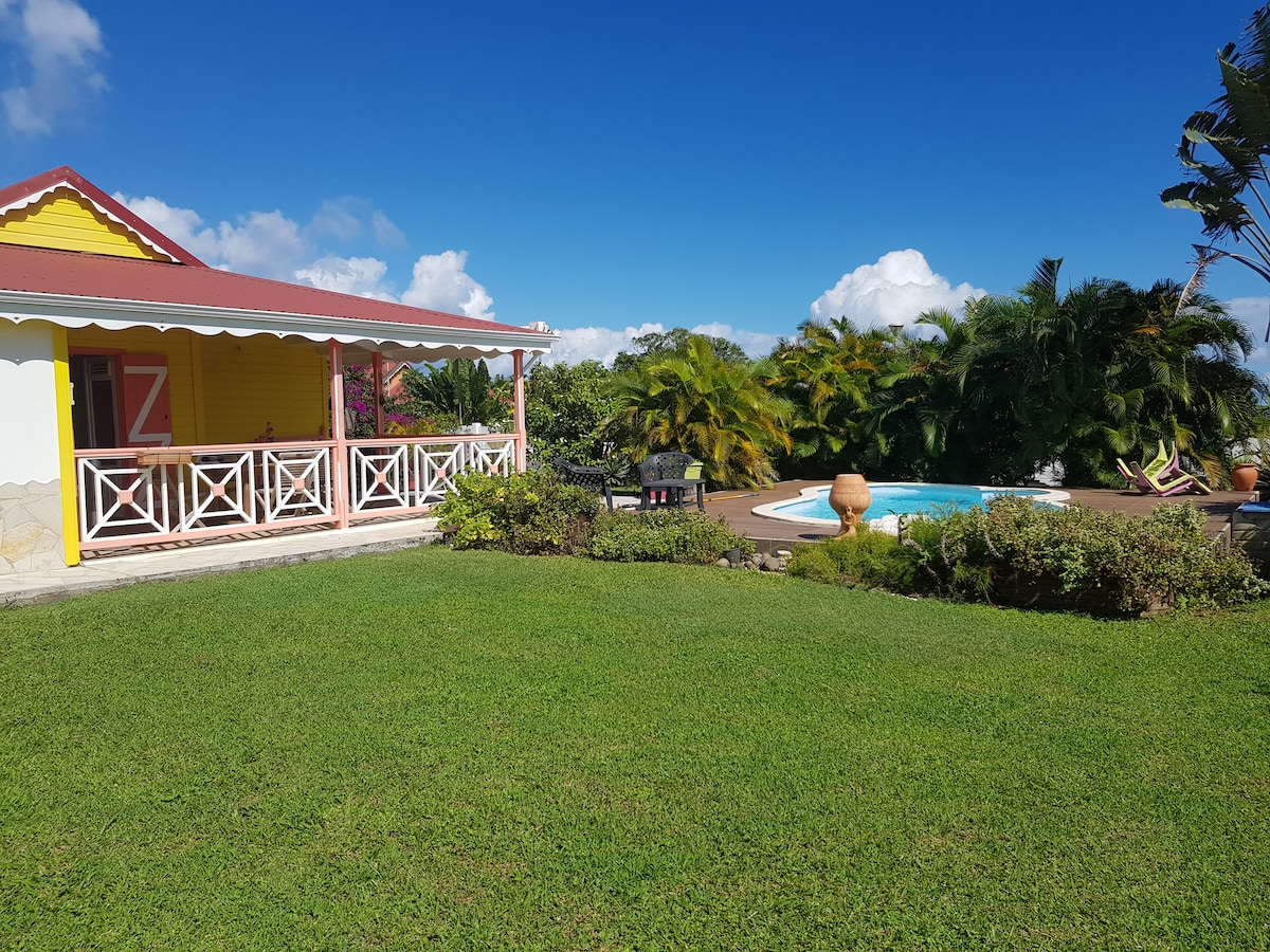 A yellow villa is set against a bright blue sky, with a well-maintained lawn leading to a private swimming pool. Lush greenery and tropical plants surround the area, contributing to a serene outdoor environment. A covered porch offers a shaded seating area.