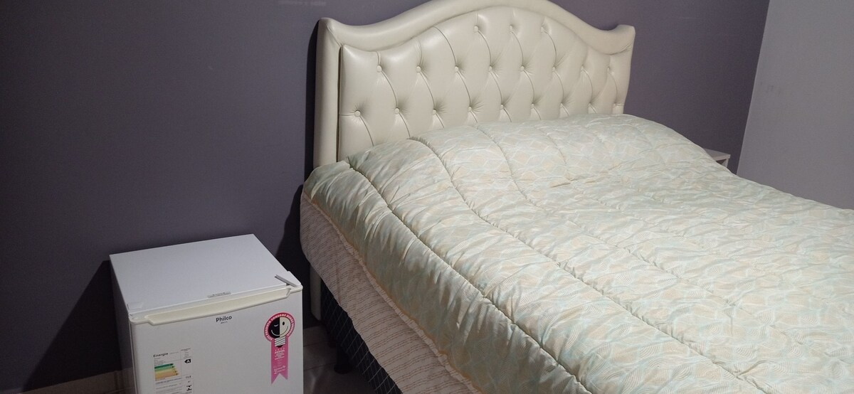 A spacious bed with a tufted, cream-colored headboard is centered against a gray wall. A light-patterned blanket covers the bed, while a compact white refrigerator is positioned next to it, offering convenience in the room.