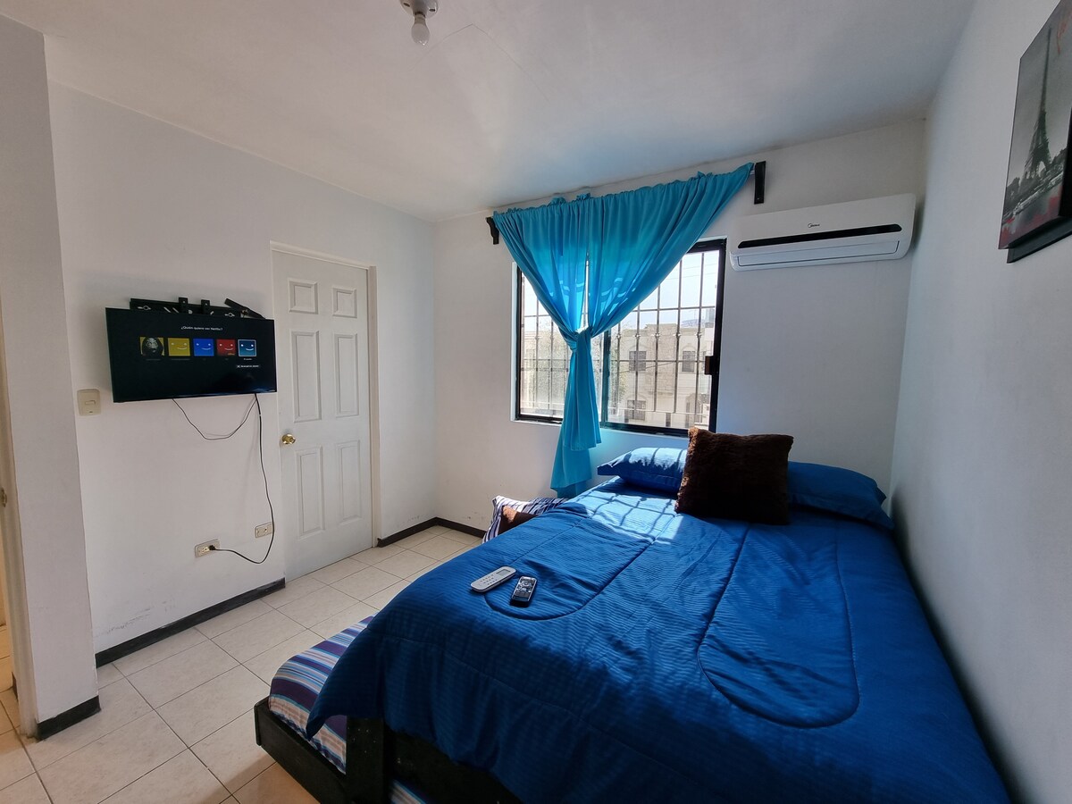 A bright bedroom features a spacious bed dressed in a blue coverlet. A window with sheer turquoise curtains allows natural light to fill the room. A wall-mounted TV is positioned opposite the bed, while a door leads to additional areas of the home.