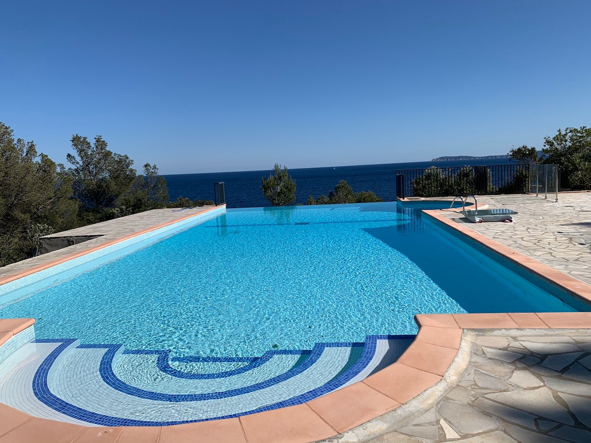 A spacious swimming pool is surrounded by natural greenery, offering panoramic views of the ocean. The clear blue water reflects the sky, and a patterned border enhances the pool's design. A sun terrace is visible, providing ample space for relaxation.