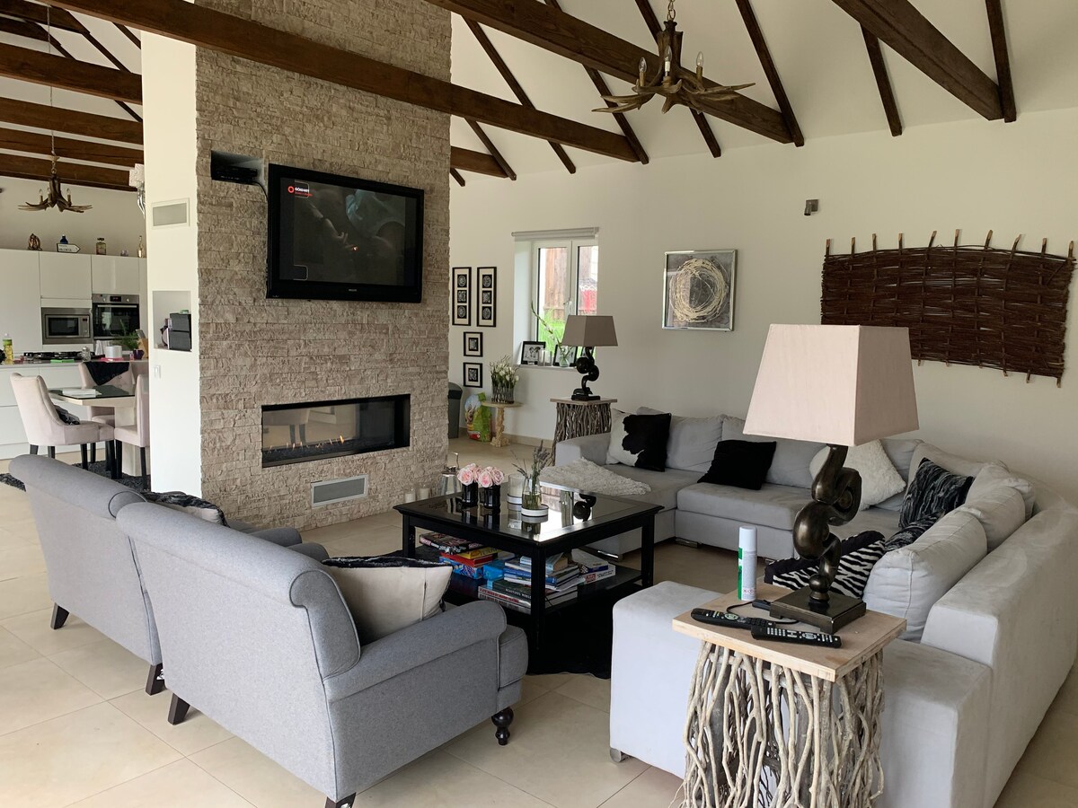 The living area features a spacious sectional sofa paired with two accent chairs, arranged around a central coffee table. A stone fireplace is mounted on the wall, with a television positioned above it. Large windows allow natural light to brighten the room.