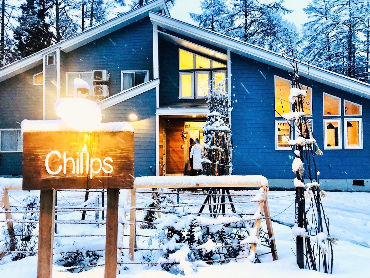 A modern blue building is surrounded by snow-covered trees, with a welcoming illuminated entrance. A wooden sign reading 'Chillps' is prominently displayed at the front, offering a warm touch to the wintry scene.