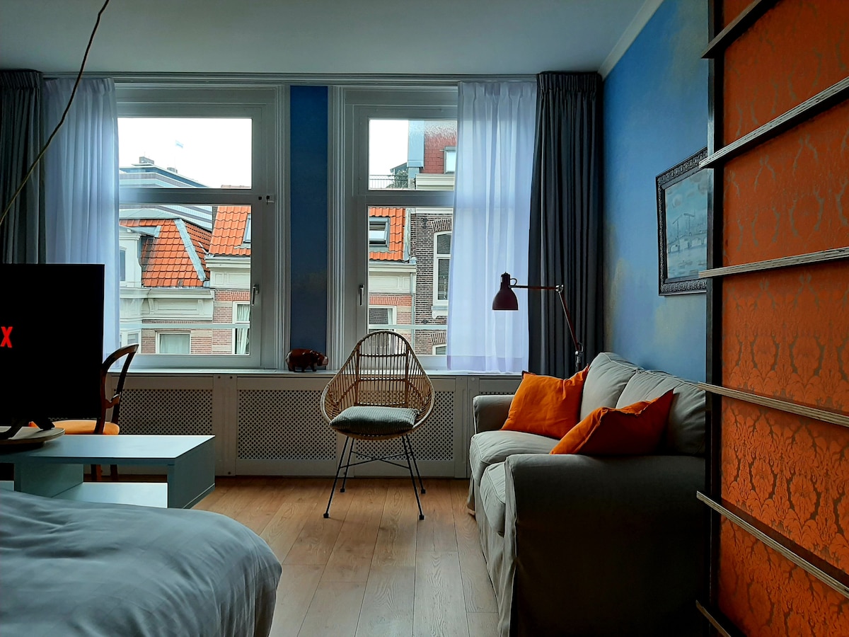 A bright and airy room is highlighted by large windows with light sheer curtains. An accent chair stands next to a cozy sofa adorned with orange cushions. A smart TV is positioned near the bed, contributing to a relaxed lounging space.