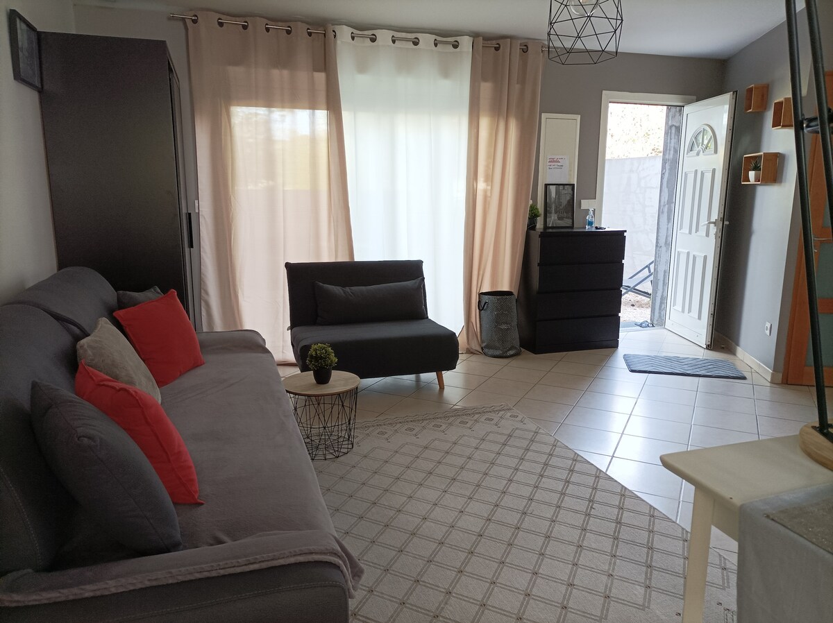 A living area is presented with a gray sofa adorned with red cushions and a matching armchair. Natural light filters through sheer curtains, illuminating the tiled floor. A dresser and a small table are visible, while a door leads to the outside.