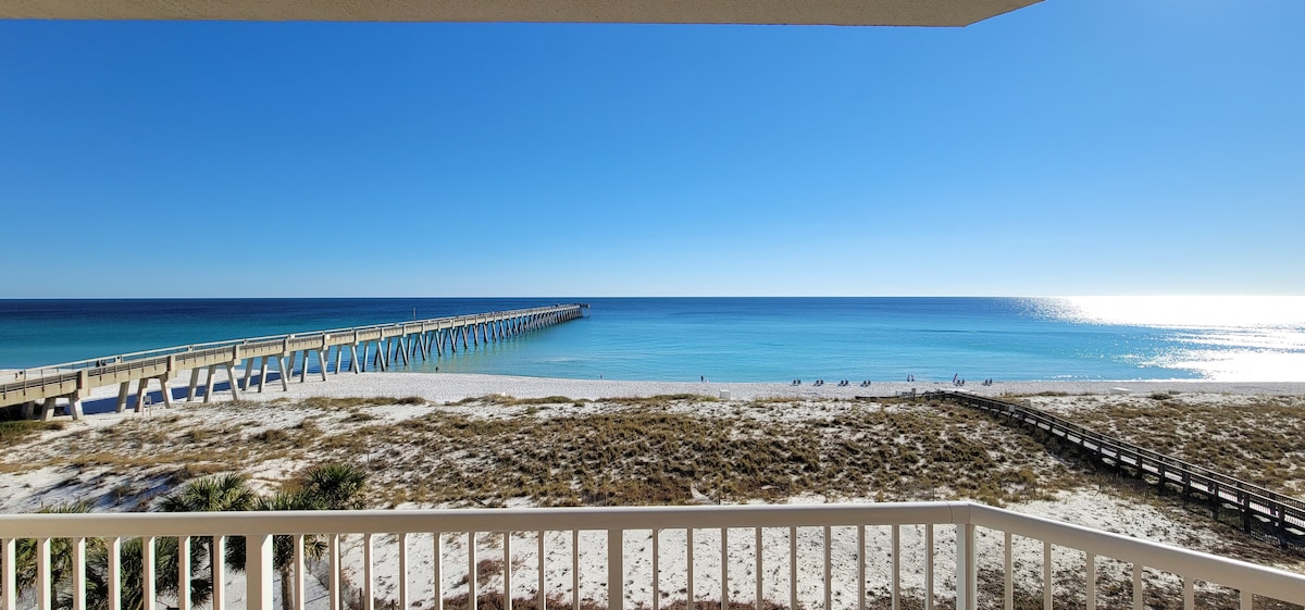 A serene view of the Gulf is presented from the balcony, highlighting the sandy beach and the Navarre Pier extending into the crystal-clear waters. The vibrant blue sky complements the tranquil landscape, inviting relaxation and enjoyment of the coastal scenery.