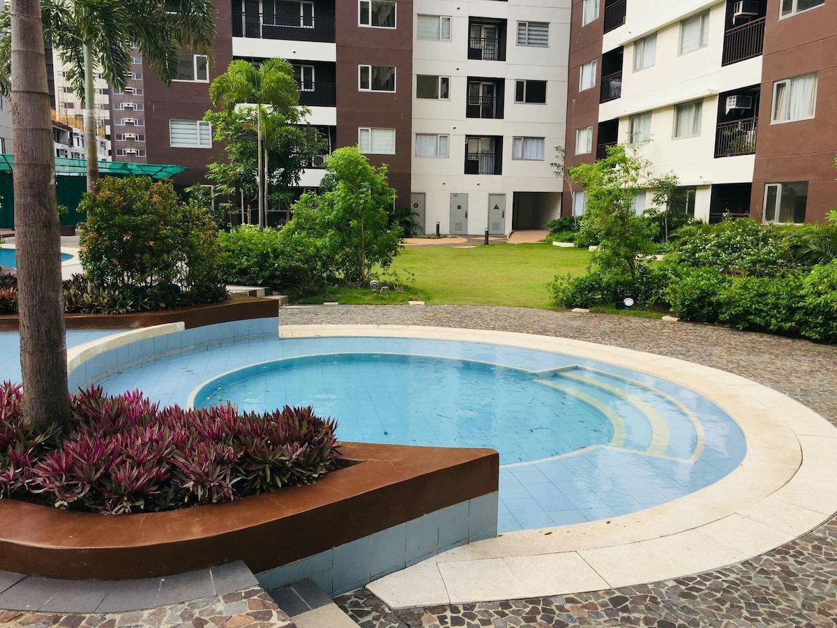 A community swimming pool is surrounded by landscaped greenery, featuring various plants and shrubs. The pool's circular shape is complemented by a small ledge for seating, creating a serene outdoor space within the complex. Nearby, well-maintained grassy areas invite relaxation and leisure.