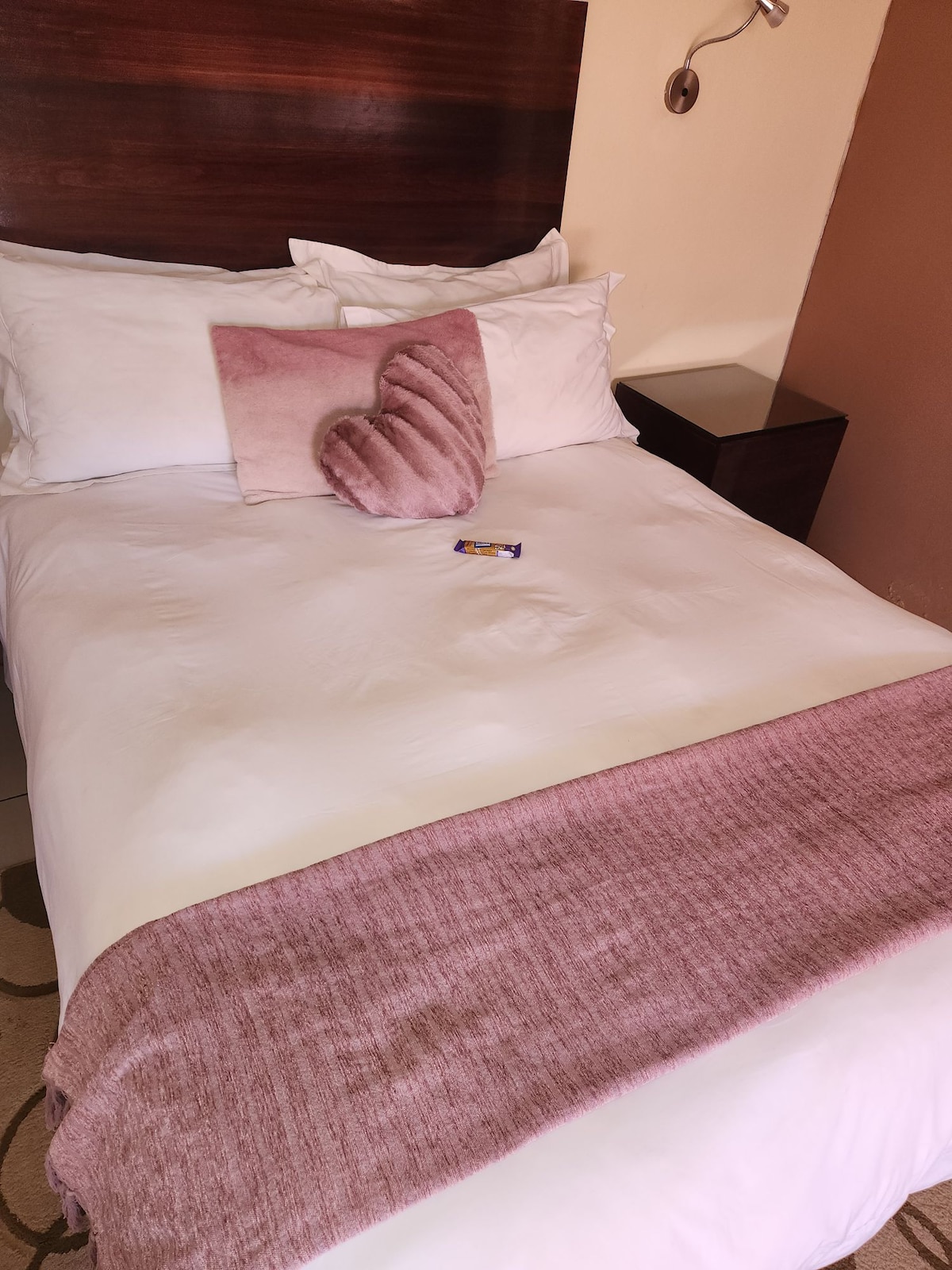 A well-appointed bed features crisp white linens and a soft pink throw blanket. A decorative heart-shaped pillow is placed atop the bed, accented by a small chocolate treat. A dark wooden nightstand is positioned next to the bed.