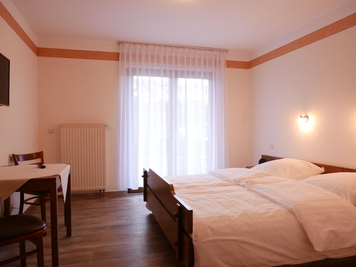 A well-lit room is presented with a spacious bed featuring fresh white linens. A small wooden table and chair are positioned nearby, while large glass doors lead to a balcony, allowing natural light to fill the space. A TV is mounted on the wall.