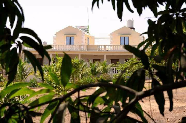A two-story retreat is partially visible through lush green foliage. The structure features a light-colored exterior with multiple windows and a balcony that overlooks the surrounding garden. Tropical plants and trees are seen in the foreground, creating a natural frame.