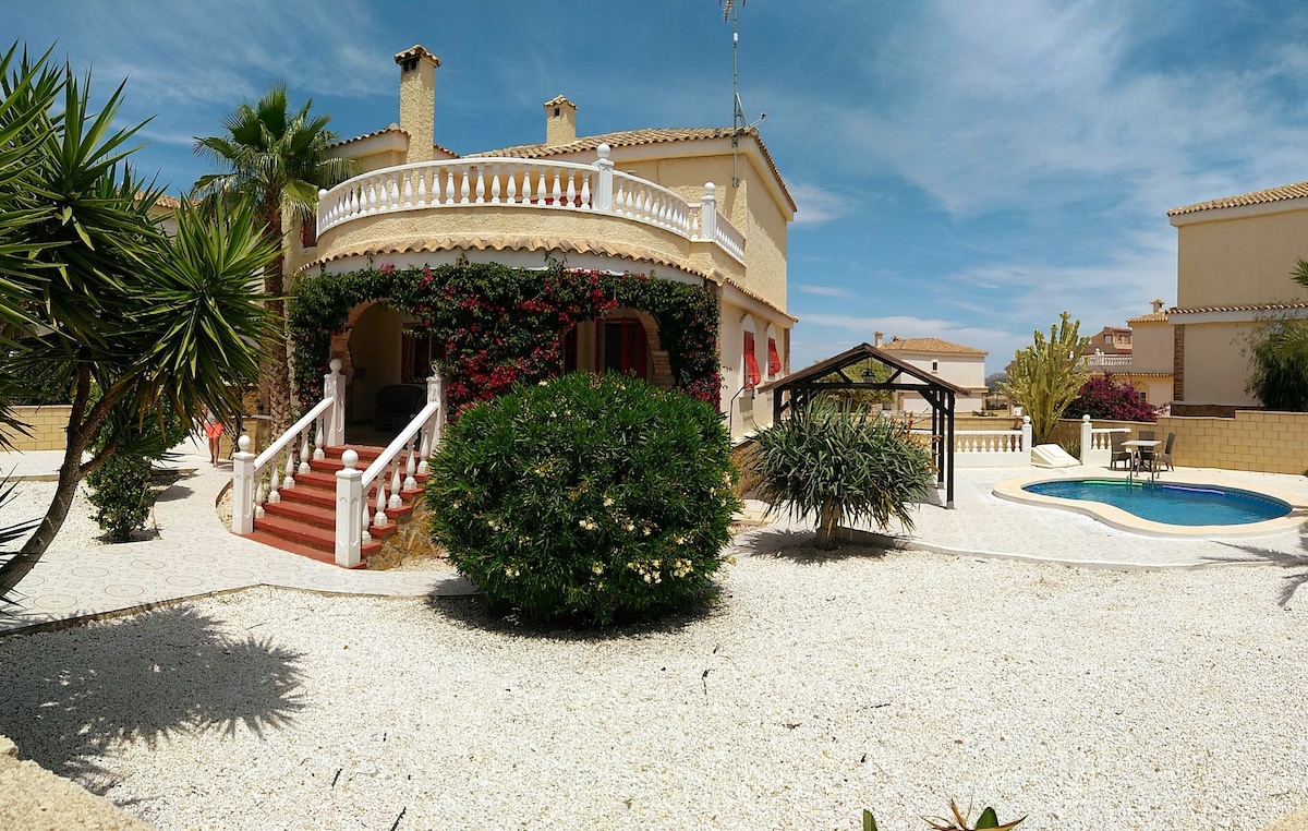 A spacious villa exterior is framed by lush greenery and colorful flowering plants. A private pool reflects the sky, while a shaded seating area is positioned nearby. The entrance features a grand staircase leading to a covered terrace, enhancing the inviting atmosphere of the home's exterior.