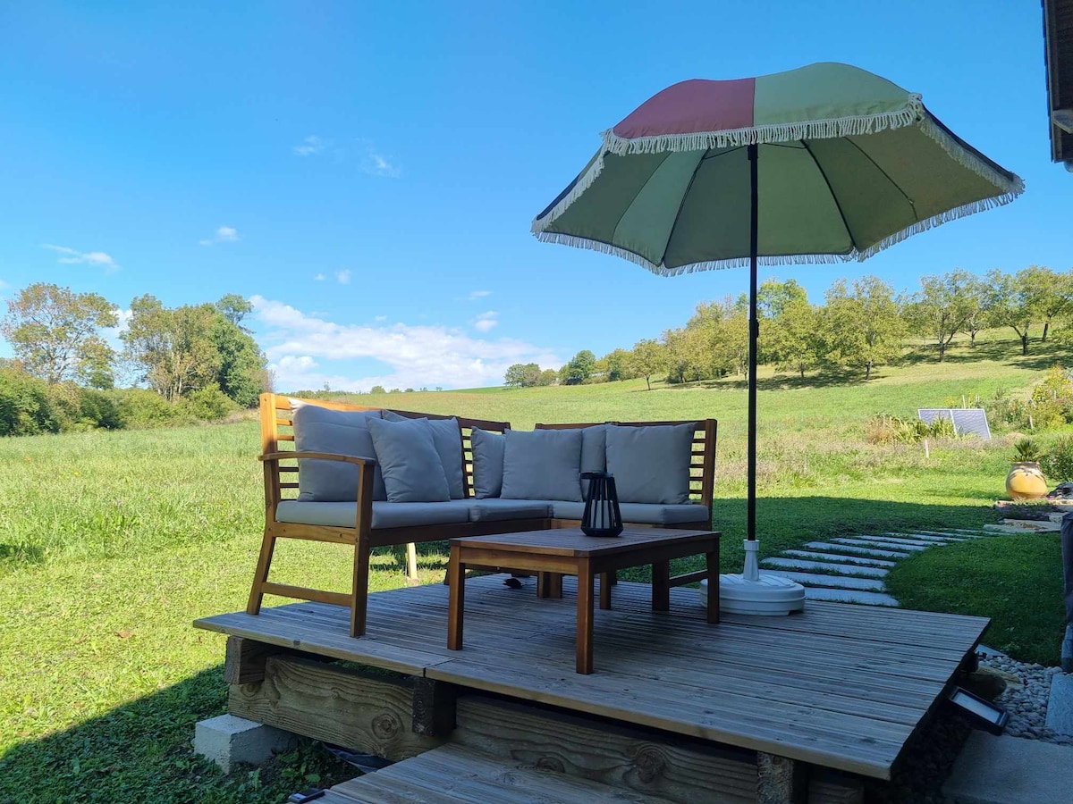 An outdoor seating area is featured on a wooden platform, accommodating a cushioned bench and a small lantern. A large umbrella provides shade, while a lush green field stretches in the background under a clear blue sky.