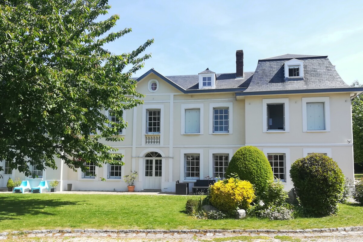 A spacious, elegant house is set in a well-kept garden, featuring large windows and a subtle color palette. The manicured lawn includes neatly shaped bushes and colorful flower beds. Outdoor seating is provided in the form of two armchairs. The atmosphere reflects a peaceful retreat.
