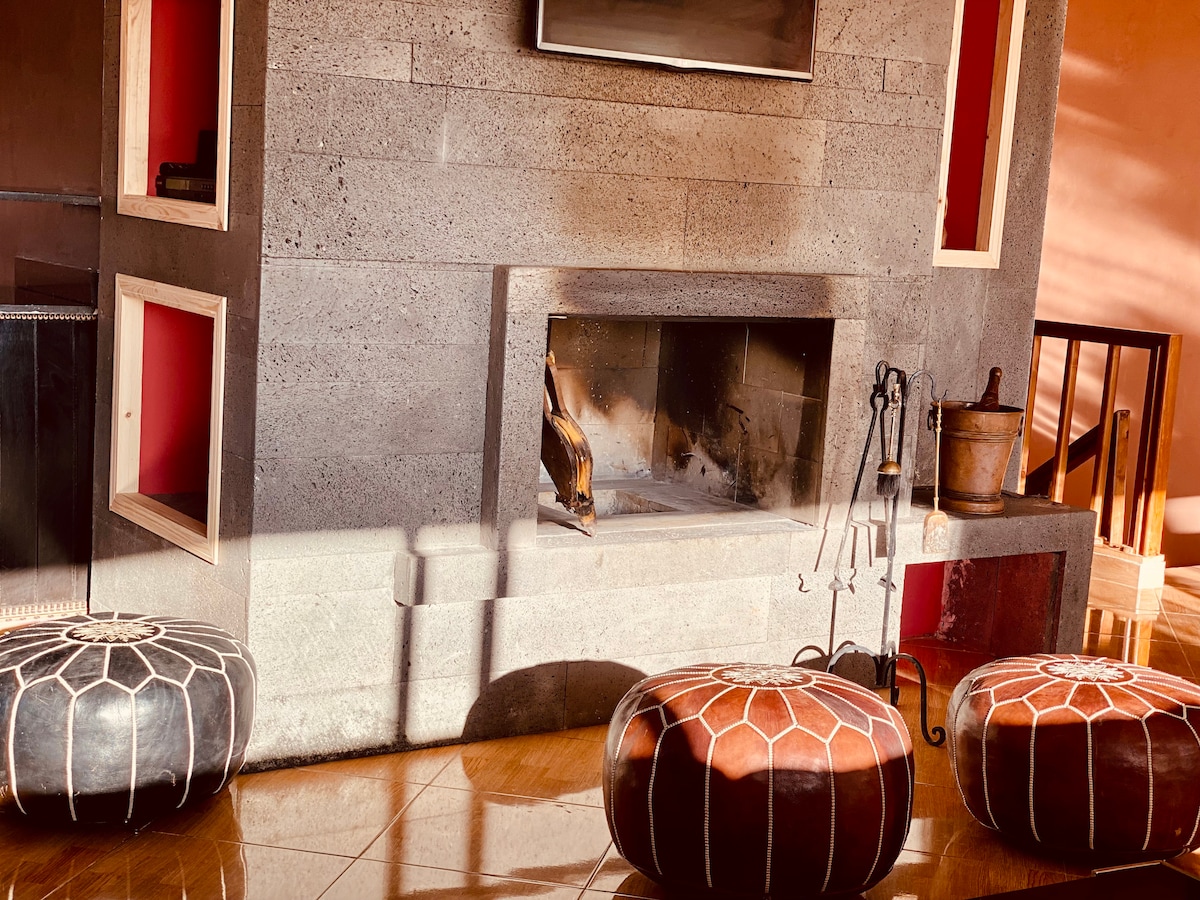 A stylish fireplace is framed by a textured stone wall, featuring a flat-screen television mounted above. Around the fireplace, round poufs in contrasting colors provide additional seating. Warm sunlight highlights the polished wooden floor, enhancing the inviting ambiance of the space.