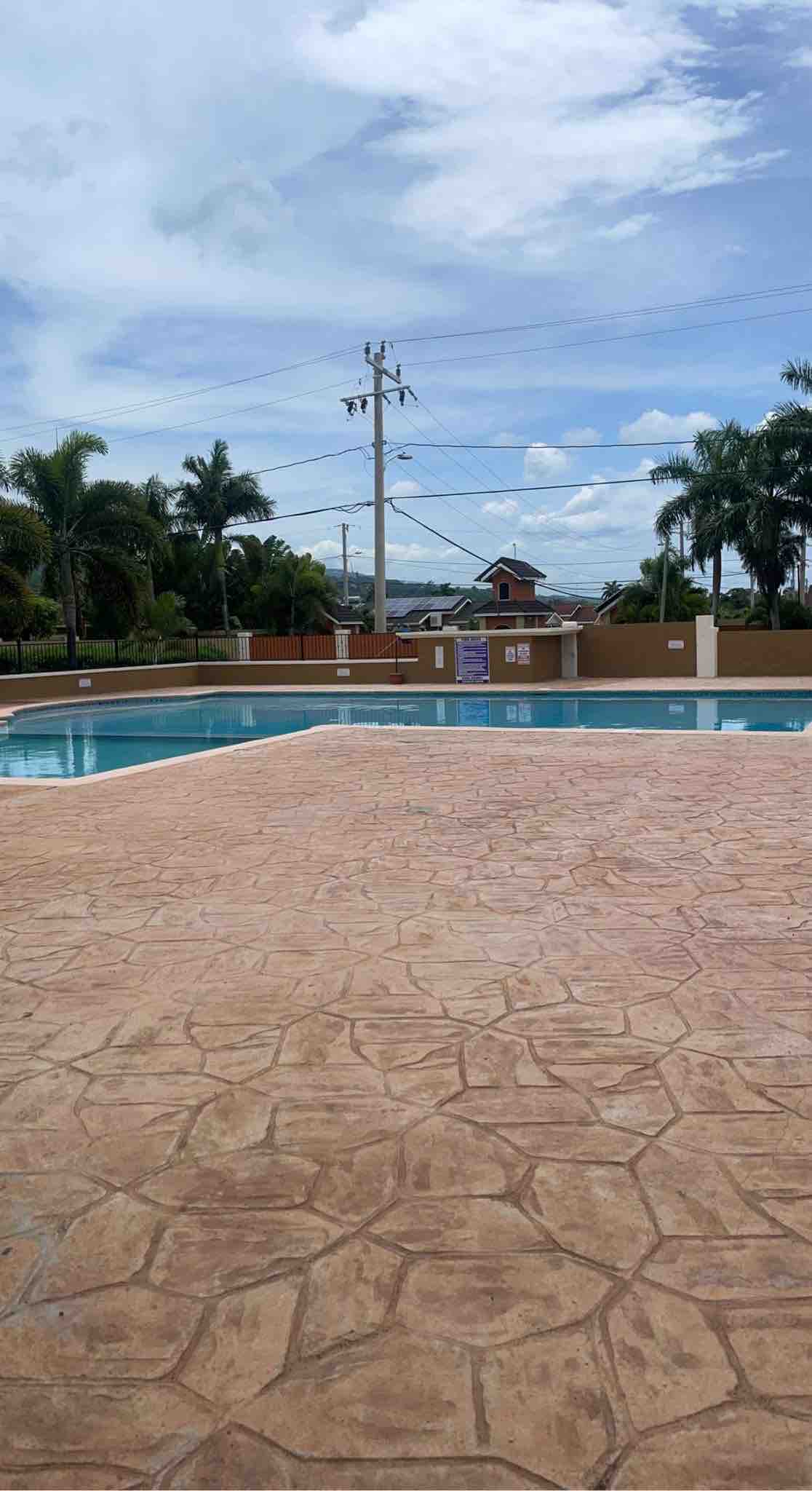 A clear view of a swimming pool is presented, surrounded by palm trees and a well-maintained patio area. The pool surface reflects the sky, with a gentle hint of blue waters. A gated community is indicated in the background, enhancing the serene atmosphere.